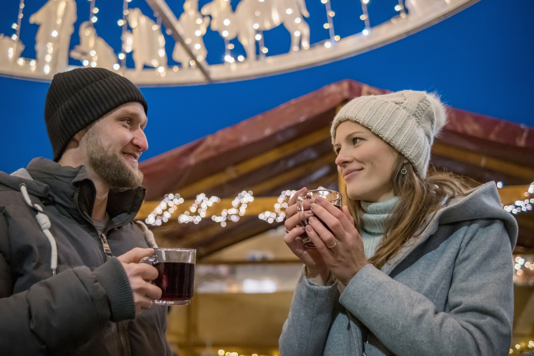 Ein Mann und eine Frau in Wintermänteln und Mützen lächeln sich an, während sie auf einem mit Lichterketten und festlichen Ornamenten geschmückten Weihnachtsmarkt Tassen in der Hand halten.